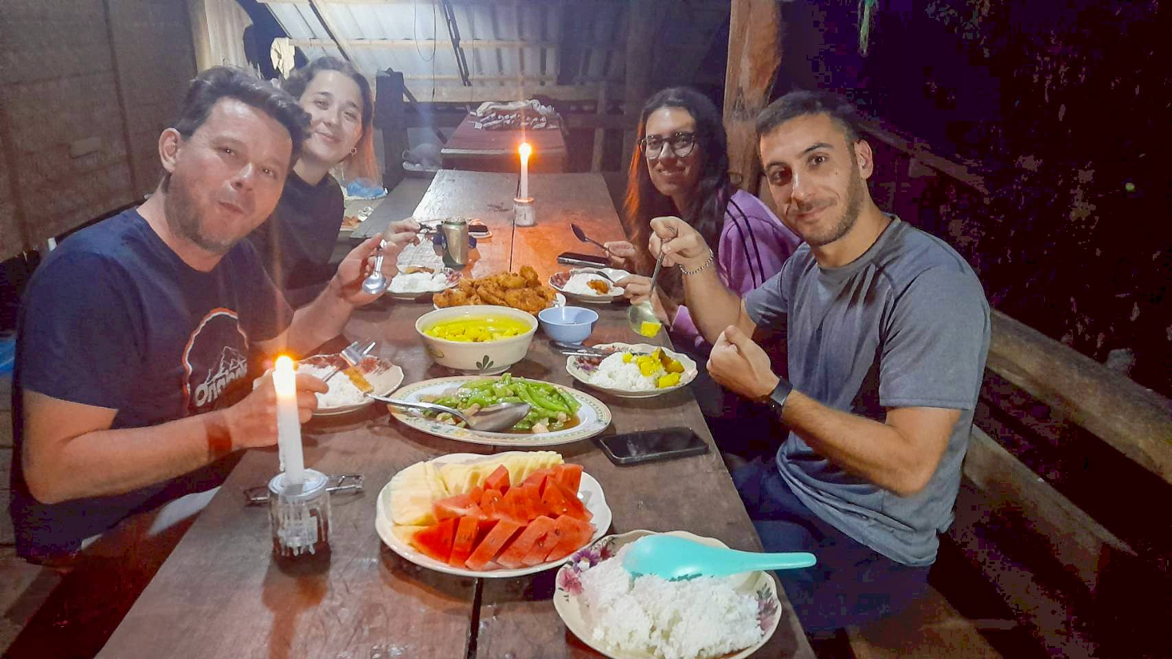 Travelers sharing a candlelit village dinner—rice, fruit, and curry on a wooden table.