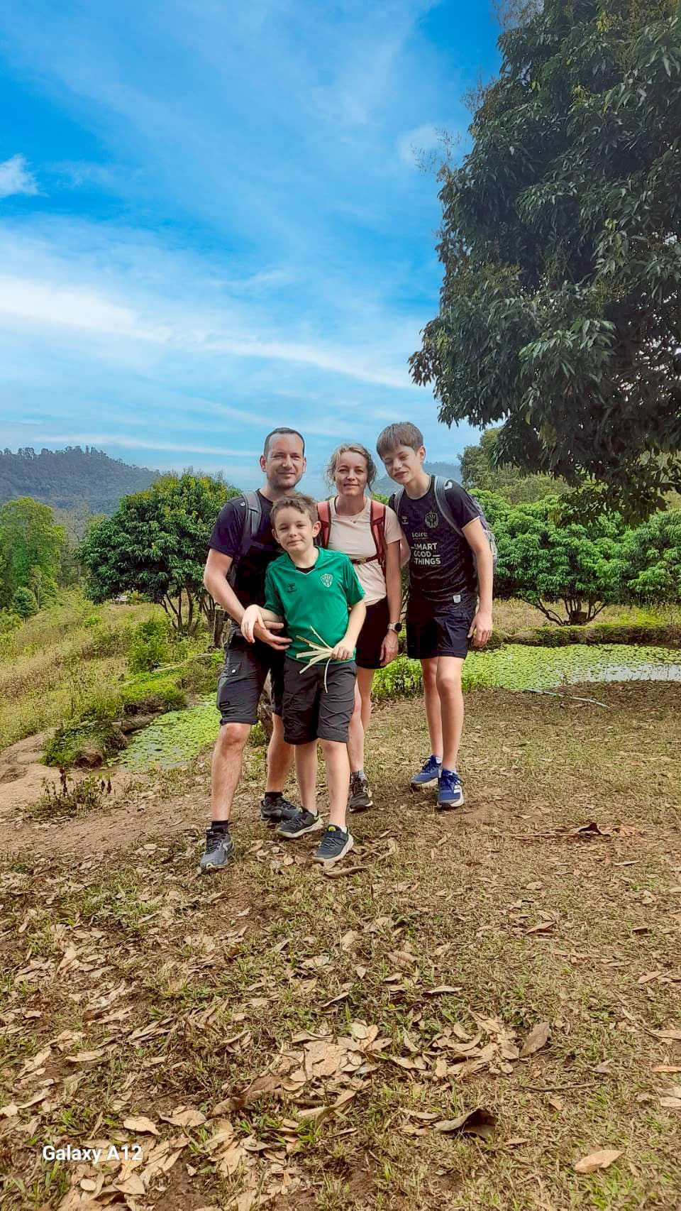 Family of four hiking on a hillside trail near Chiang Mai, smiling under a blue sky.